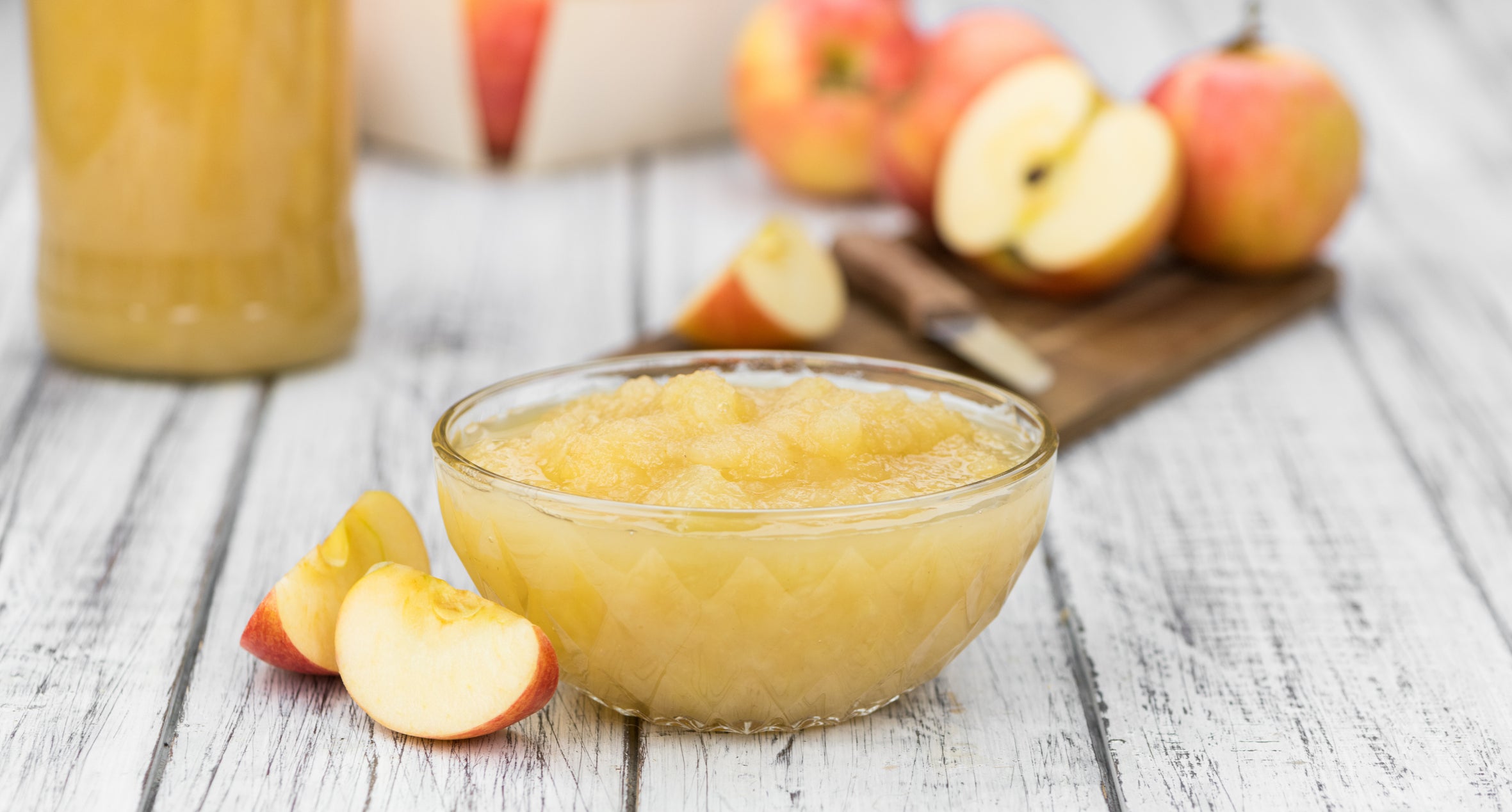 Glass bowl of applesauce with sliced apples and a jar in the background on a wooden surface