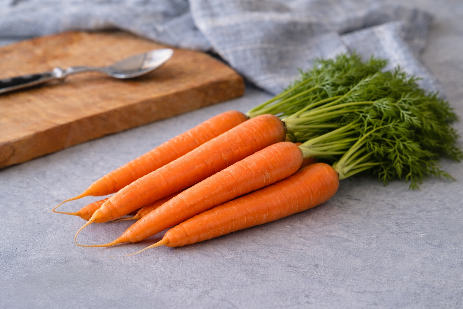Bunch of carrots on a gray surface with a wooden cutting board and spoon in the background.