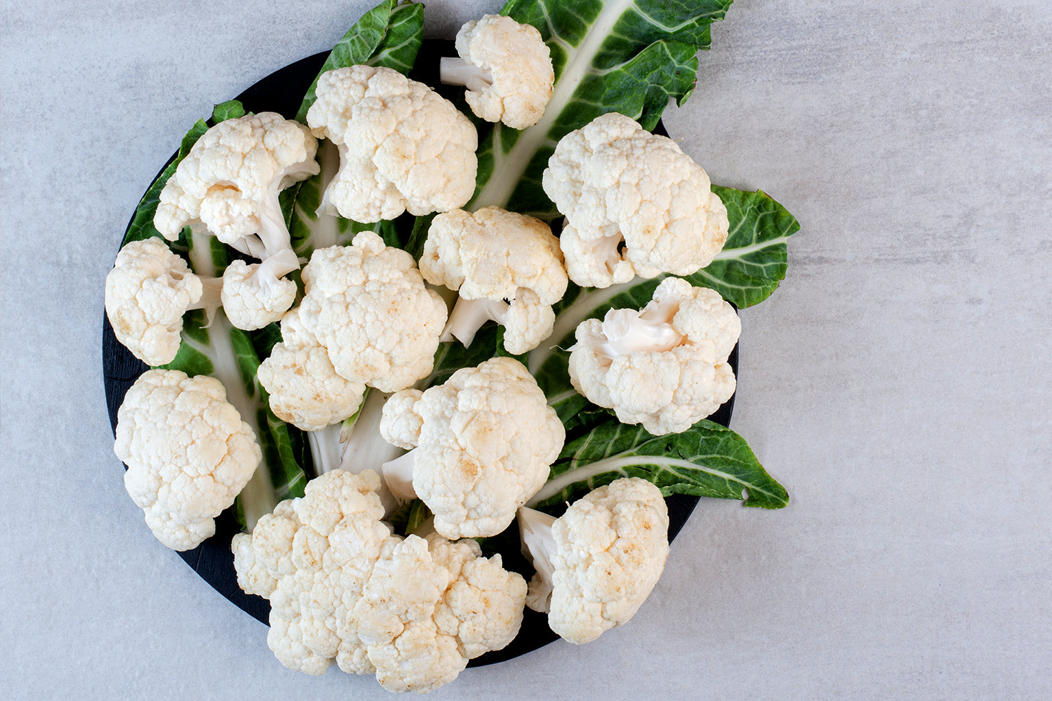 White cauliflower florets on a dark plate with green leaves on a light gray background