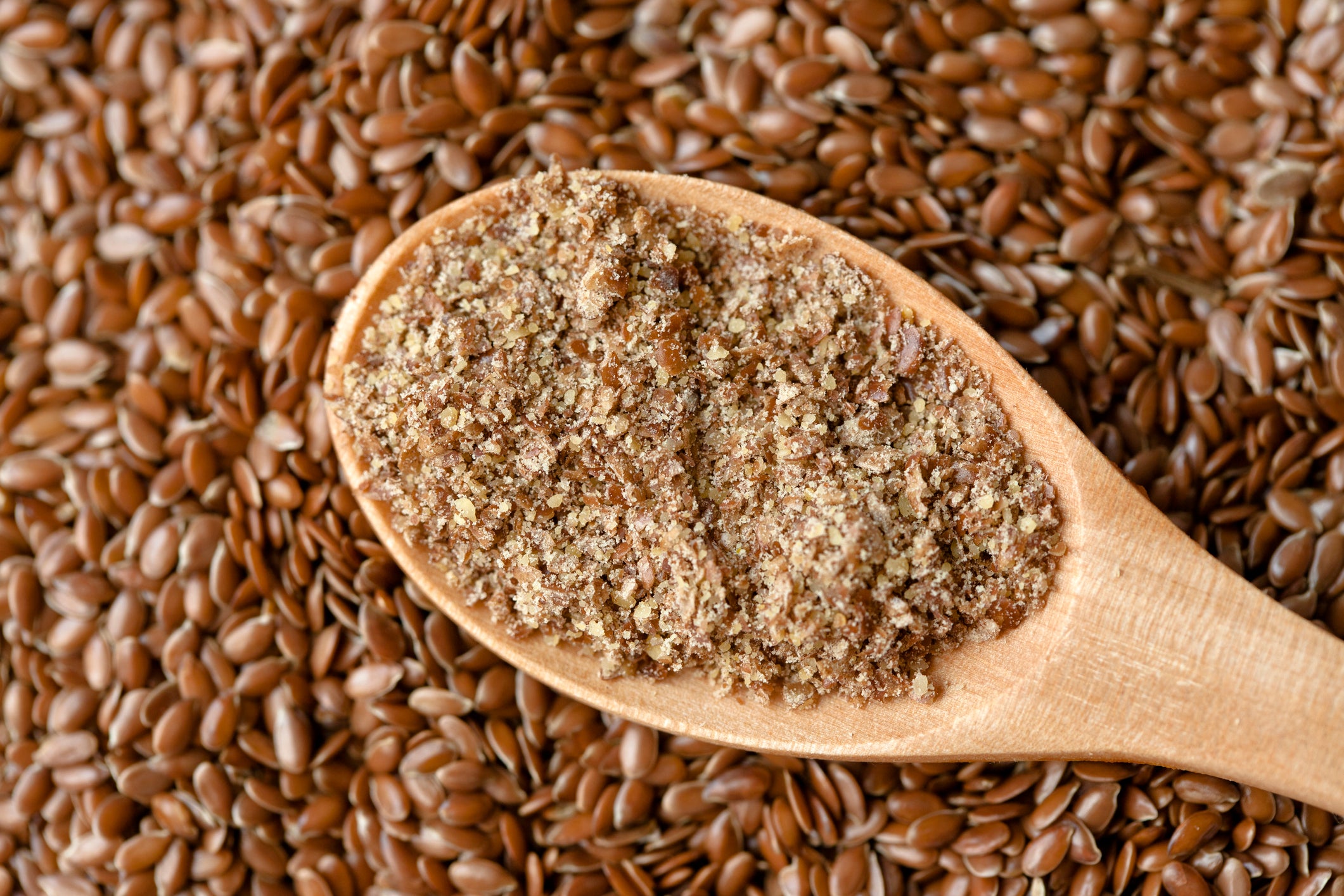 Wooden spoon filled with ground flaxseed surrounded by whole flaxseeds.