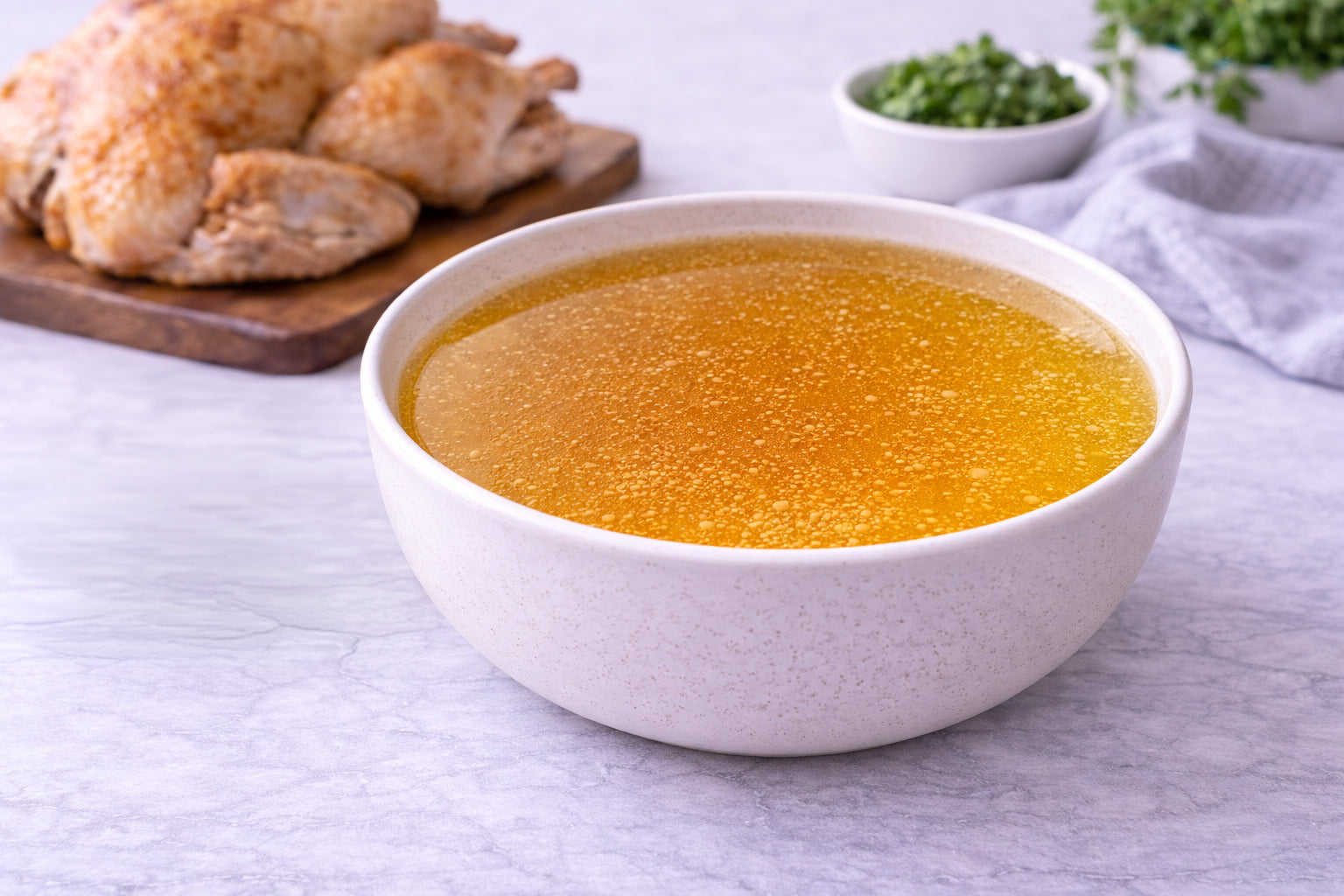 Bowl of chicken broth with roasted chicken pieces and herbs on a table.