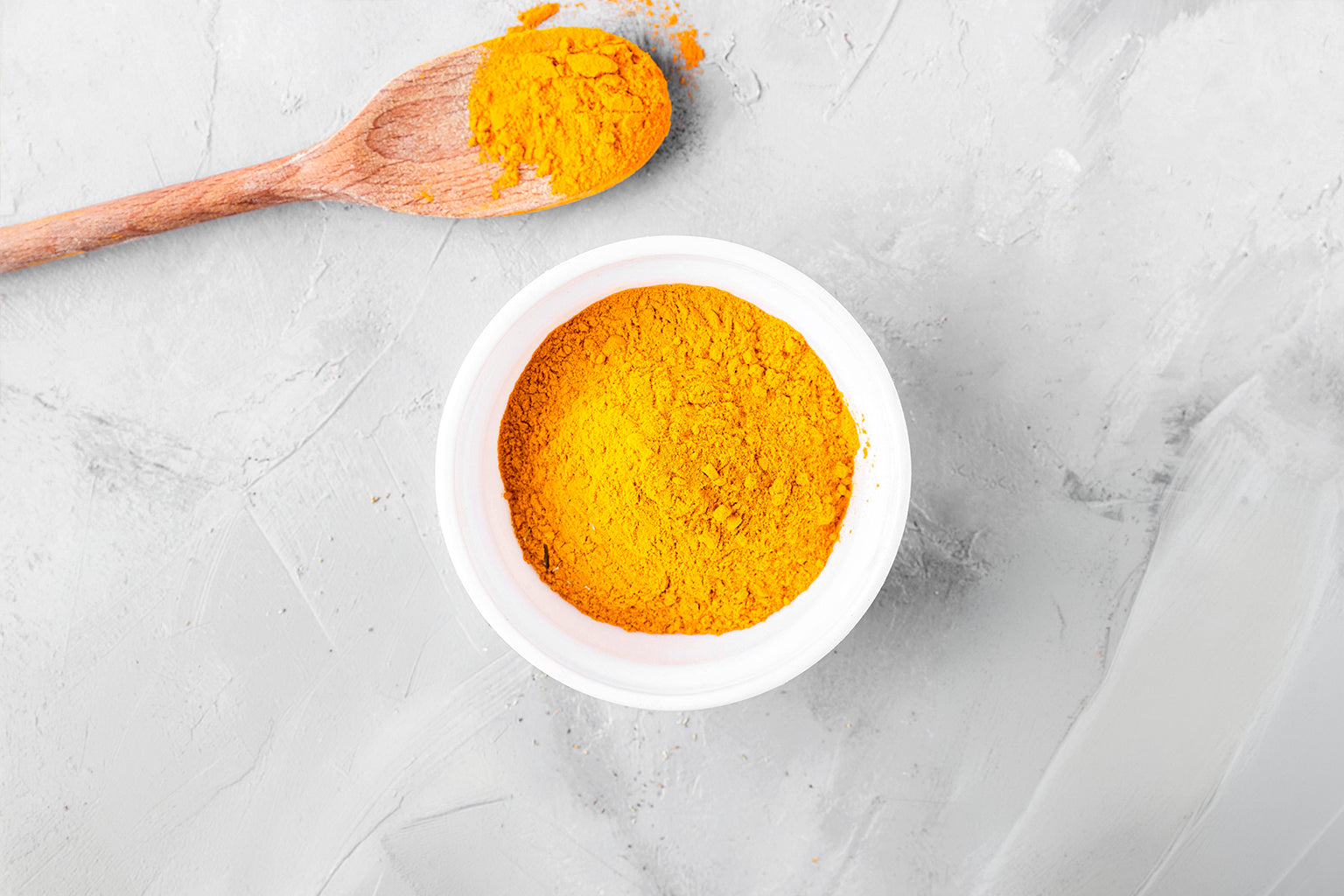 Turmeric powder in a white bowl with a wooden spoon on a light gray background