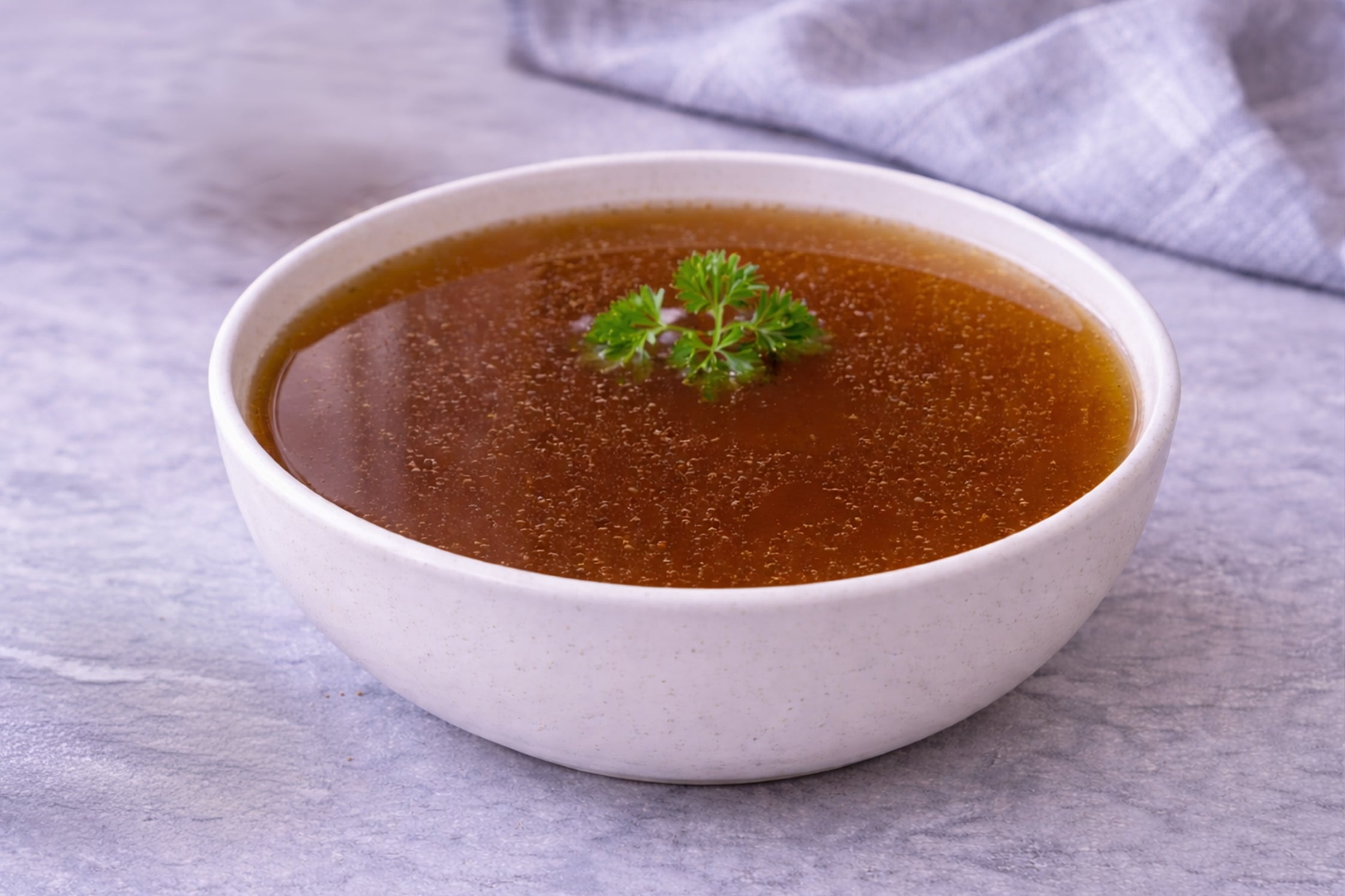 Bowl of beef broth with a sprig of parsley on a gray surface